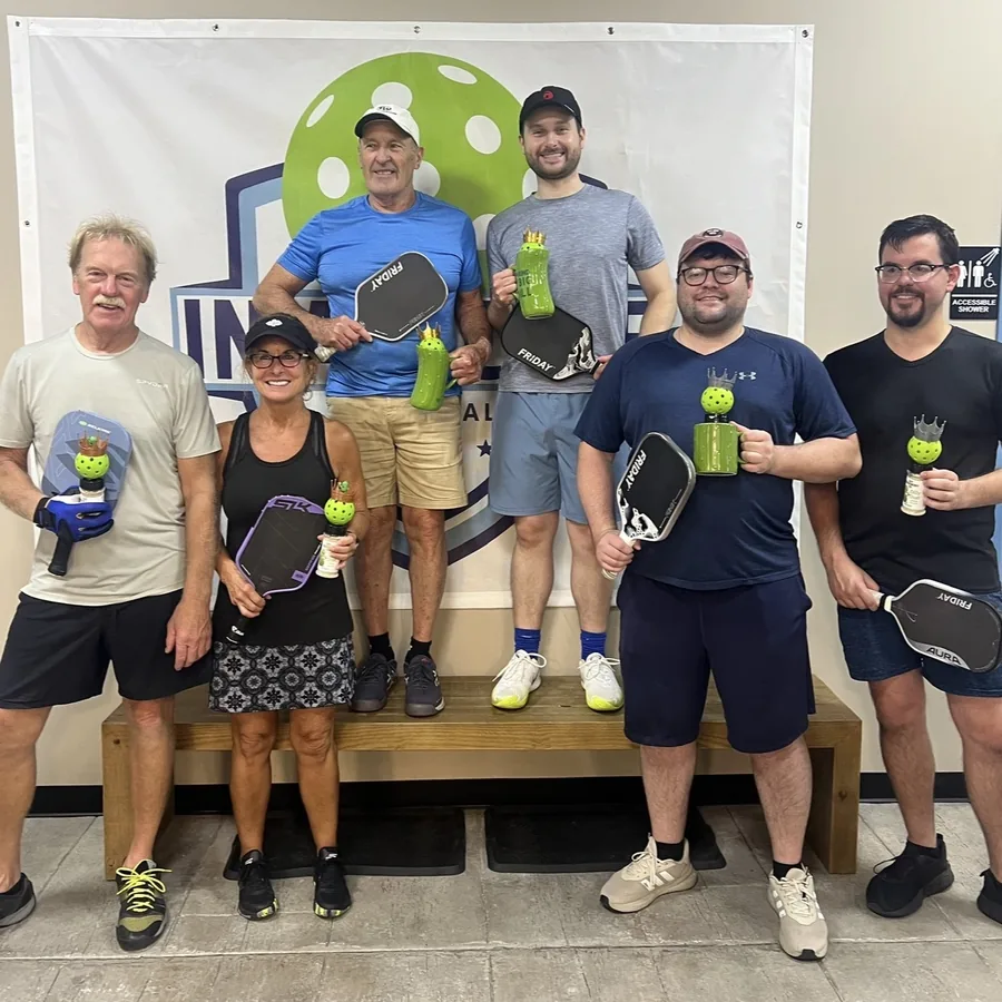 Six players holding paddles and pickleball trophies in front of the club banner